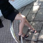Woman in a black dress seated on a metal railing with legs crossed, wearing black high-heeled sandals; sunlit sidewalk and tiled wall behind.