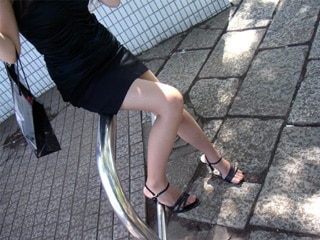 Woman in a black dress seated on a metal railing with legs crossed, wearing black high-heeled sandals; sunlit sidewalk and tiled wall behind.